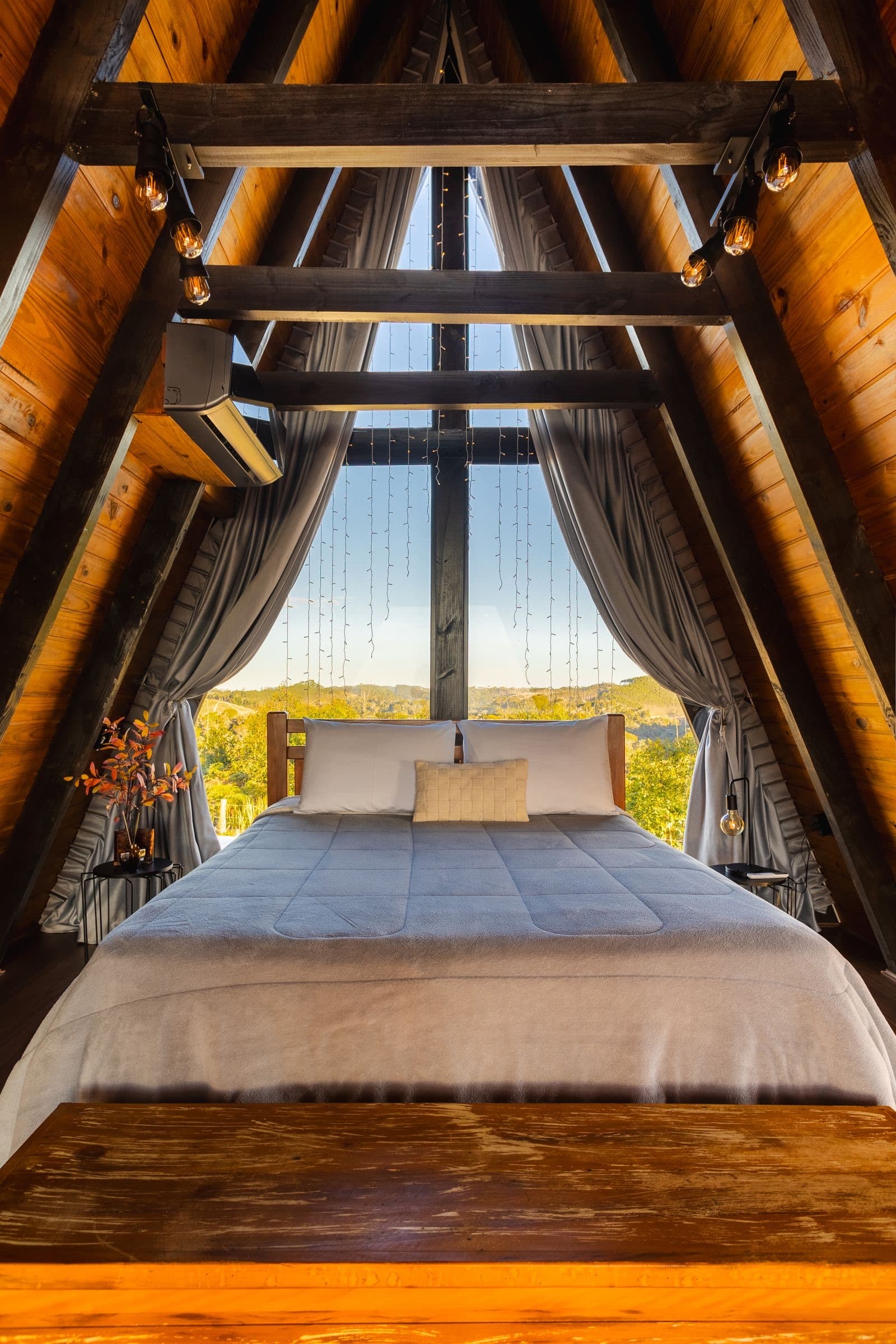 A loft bedroom under a timber A-frame with a window onto the hills
